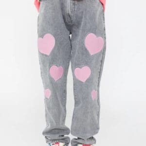 A person wearing (Made Extreme)M (10) grey jeans with pink heart patches, a pink t-shirt with a white graphic, and white sneakers stands against a plain white background.