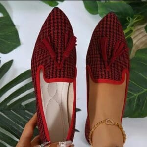 A pair of Houndstooth Flats With Tassle—one in hand, the other worn with an anklet—set against a leafy backdrop.