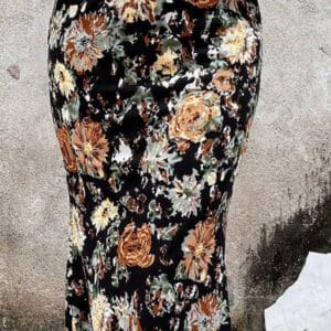A person wearing a white long-sleeve top and the Flower Skirt—a high-waisted, black floral print skirt—stands against a textured, light gray wall.
