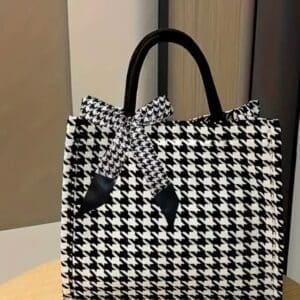 The Houndstooth Purse, featuring a black and white houndstooth pattern with black handles and a matching bow, sits on a wooden surface.
