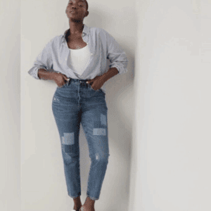 A person stands against a white wall wearing a striped button-up shirt over a white top, Dark Denim Jeans with denim patches and white stitching, and flats.