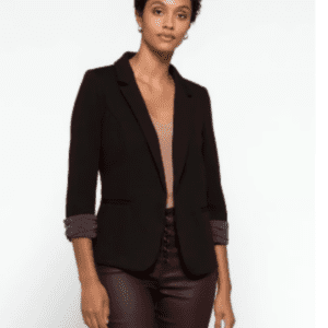 A person with short curly hair wearing a black blazer over a brown top and burgundy pants stands against a plain white background.