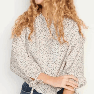 A person with long curly hair, smiling and looking to the side, is wearing the Mini Flowers Top—a loose, light-colored blouse with a floral pattern—paired with blue jeans.