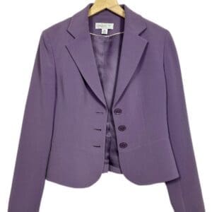The Cropped Pleated Back Blazer in purple, featuring a notched lapel and three-button closure, hangs on a wooden hanger against a white background.