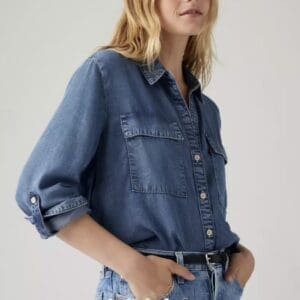 A person wearing a long-sleeve denim shirt with front pockets and rolled-up sleeves, paired with light blue denim jeans and a black belt.