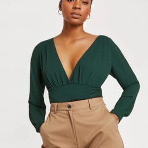 A woman stands with her hands in her pockets against a plain white background, wearing a deep green Long Sleeve Low Cut Bodysuit and high-waisted tan pants.