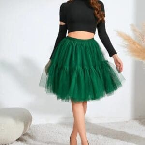 A woman poses indoors wearing a black long-sleeve crop top, a green layered tulle skirt, and white heels on a white carpeted floor.