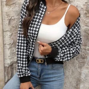 A woman wearing a black and white houndstooth jacket over a white tank top and jeans holds a coffee cup while standing against a textured wall.