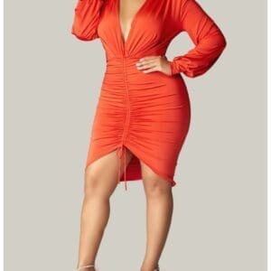A woman poses in a long-sleeve, deep V-neck, red ruched dress with an asymmetrical hem, paired with silver high-heeled sandals against a plain background.