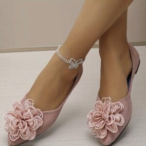 A pair of feet wearing pink flat shoes with large fabric flower decorations and a sparkly anklet featuring a butterfly design.