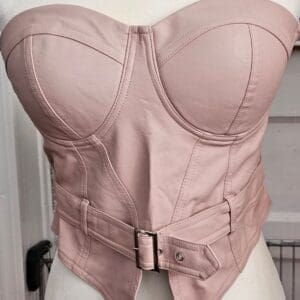 A blush pink strapless leather corset with structured cups, visible seams, and a matching belt with a silver buckle displayed on a mannequin.