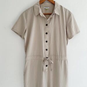 A beige short-sleeve button-up jumpsuit with a collar and waist tie, hanging on a wooden hanger against a white background.