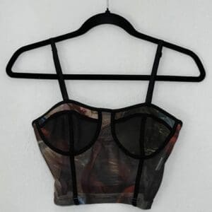 A cropped tank top with black straps and trim, featuring a muted print hanging on a black hanger against a plain white wall.