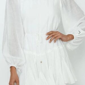 A woman wearing a long-sleeved, white, ruffled mini dress with a cinched waist, standing against a plain light background.