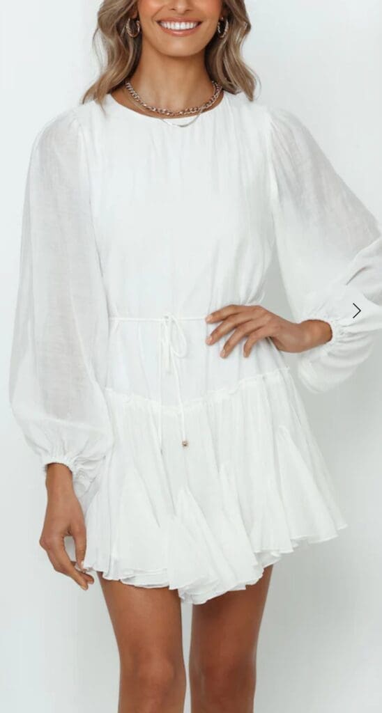A woman wearing a long-sleeved, white, ruffled mini dress with a cinched waist, standing against a plain light background.