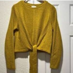 A mustard yellow, long-sleeve knit sweater with a front tie, hanging on a hanger against a white door.