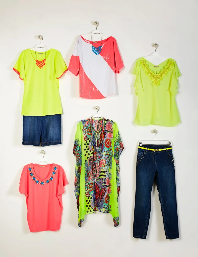 Six colorful women's outfits including tops, shorts, a kaftan, and jeans are displayed on hangers against a plain white background.