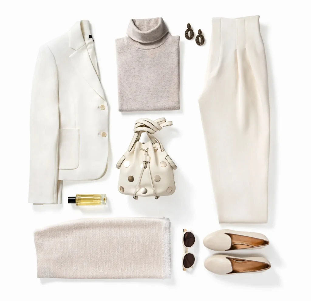 A flat lay of cream and beige clothing and accessories, including a blazer, turtleneck, trousers, skirt, purse, shoes, sunglasses, earrings, and a perfume bottle.