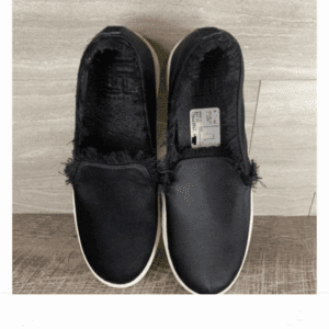 A pair of black slip-on shoes with frayed edges and white soles placed side by side on a wooden surface. A size label is visible inside the right shoe.