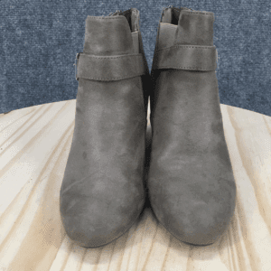 A pair of gray suede ankle boots with side zippers and strap details, positioned upright on a light wooden surface against a textured gray background.