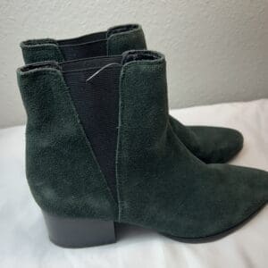 The Green Geel Suede Bootie features dark green suede, black elastic side panels, and chunky low heels. Displayed on a white surface with a light-colored wall in the background.