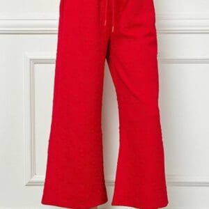 Wearing Red Heart Crop Pants—bright red, textured, with a wide-leg fit, elastic waistband, and drawstring—a person pairs them with white sneakers while standing against a white paneled wall.