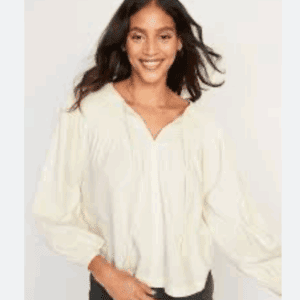 Woman wearing a loose, long-sleeved, cream-colored blouse and dark pants, standing and smiling at the camera against a plain background.