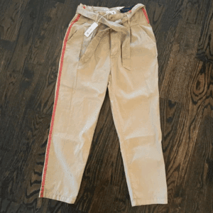Khaki paperbag waist pants with a fabric belt, red side stripes, and front pockets, displayed flat on a dark wooden floor. Tags are attached to the waistband.
