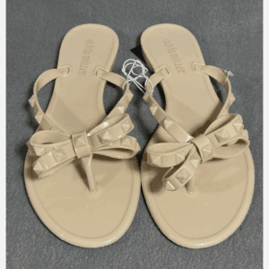 A pair of beige flat sandals with decorative bows and stud accents on the straps, displayed on a gray surface.