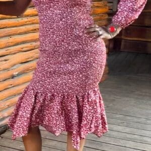 Woman posing on a wooden porch wears the One Shoulder Qtr Sleeve Gathered Dress in pink and white floral, paired with tan lace-up heels and red accessories.