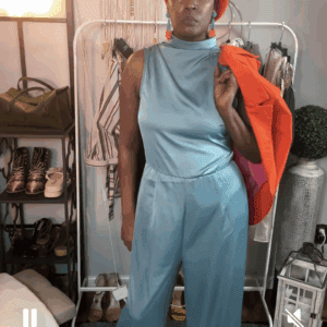 A woman stands indoors wearing a Turquoise Sleeveless Satin Jumpsuit, an orange beret, and holding an orange jacket over her shoulder. Clothing and accessories can be seen in the background.