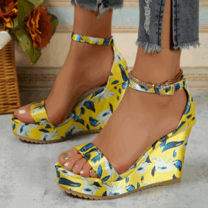 A person wearing yellow floral platform wedge sandals and frayed-hem jeans stands on a light-colored floor with a wicker basket and orange flowers in the background.