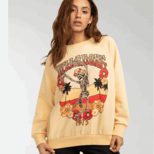 Person wearing a pale yellow Billabong sweatshirt with a graphic of a skeleton, flowers, palm trees, and "1973" printed on the front.