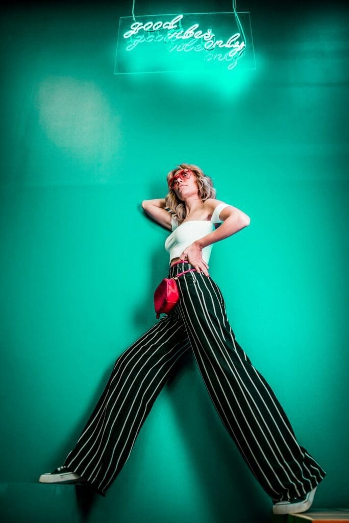 A woman poses against a teal wall wearing striped wide-leg pants and a white top, with a neon sign reading "good vibes only" glowing above her.