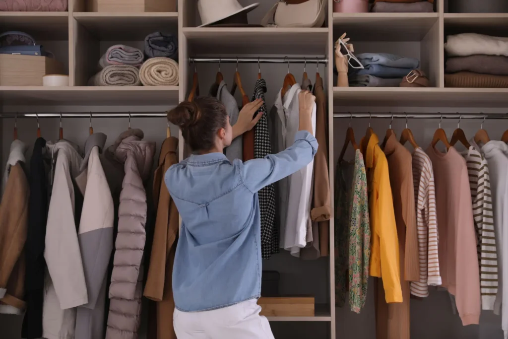 A woman stands in front of an organized closet, reaching for a shirt among neatly hung clothes and folded items on shelves.