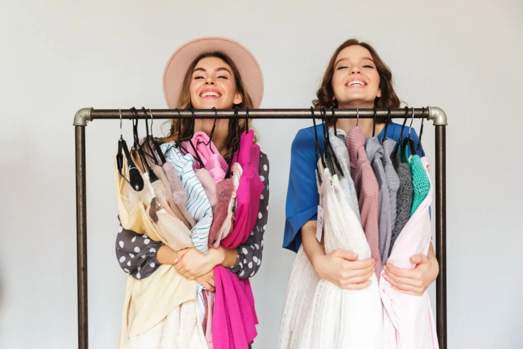 Two women stand behind a clothing rack, smiling and holding various colorful garments draped over hangers.