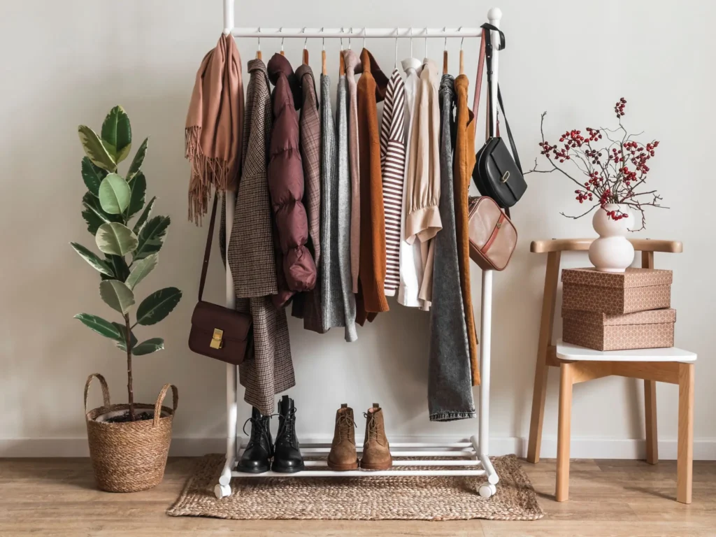 A white clothing rack holds various jackets, tops, and bags. Boots sit below. Nearby are a potted plant, stacked boxes on a chair, and a vase with red berries.