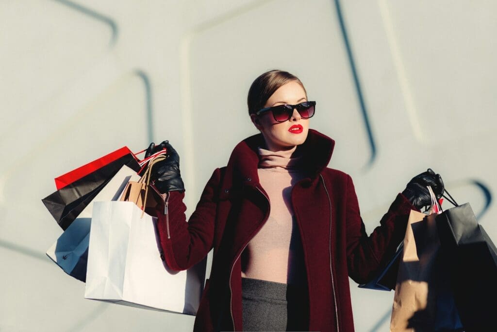 A woman wearing sunglasses and a dark coat holds multiple shopping bags in both hands, standing against a light-colored, modern wall.