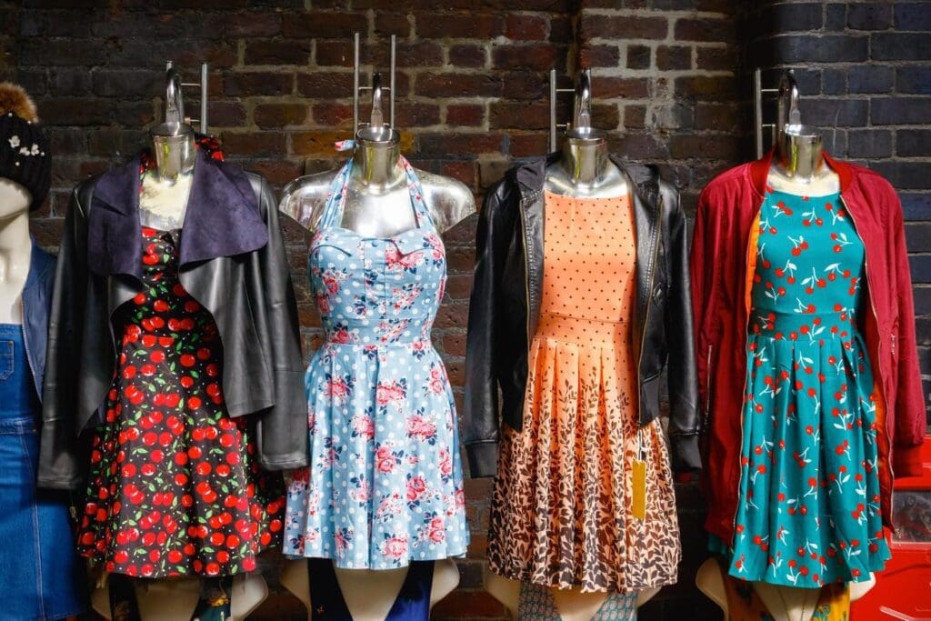 Four mannequins display colorful dresses and jackets against a brick wall, each featuring different patterns, including floral and polka dots.