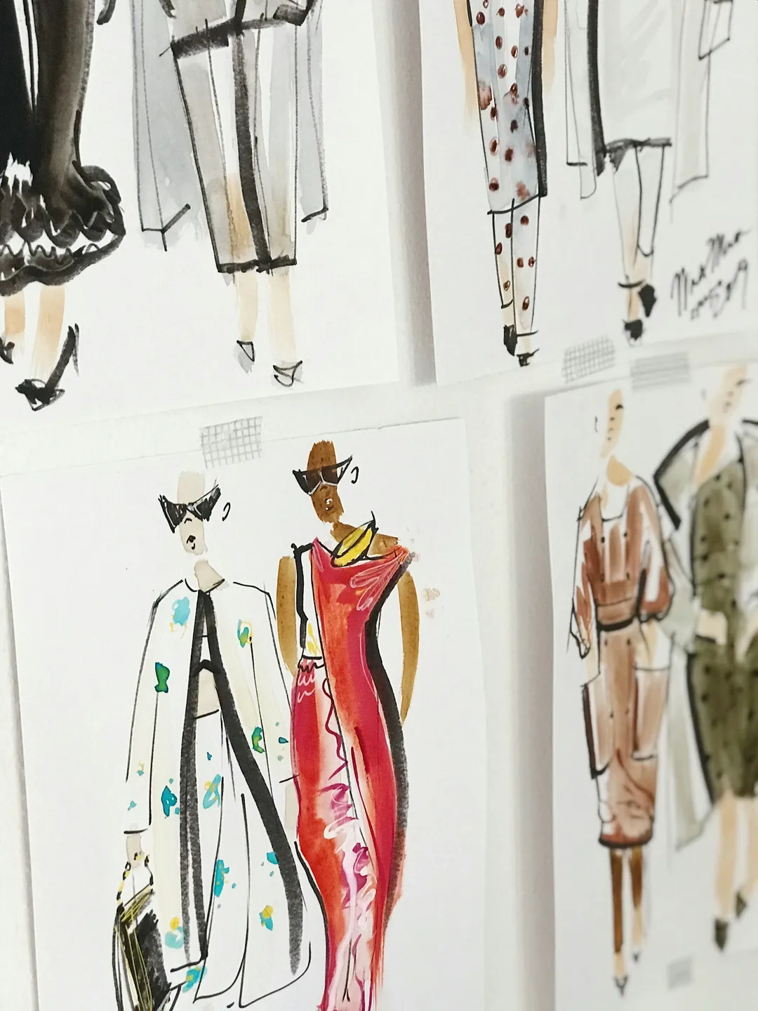 Fashion sketches of various women’s outfits are pinned to a wall, featuring colorful and patterned clothing designs in watercolor and ink.