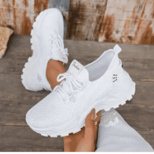 A person wearing white athletic sneakers with textured soles, sitting on a wooden surface. The shoes have laces and a mesh-like upper design.