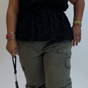 A person wearing olive green Cargo Jogger pants, a black short-sleeve blouse, black shoes, and colorful beaded bracelets holds a black item with a strap.