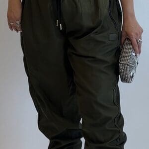 Person wearing a black graphic T-shirt, Smooth Parachute Joggers in olive green, strappy beige heels, and holding a silver clutch, standing against a plain background.