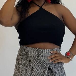 Person wearing a sleeveless black halter top and gray patterned skirt, accessorized with red earrings, a red ring, and a bracelet, posing against a plain background.
