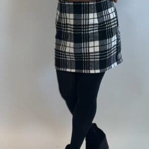 Person wearing a black and white plaid skirt, black tights, and black shoes, standing against a plain light-colored background.