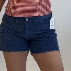 A person wearing blue denim shorts with a tag and a red-and-white checkered top, standing against a plain background.