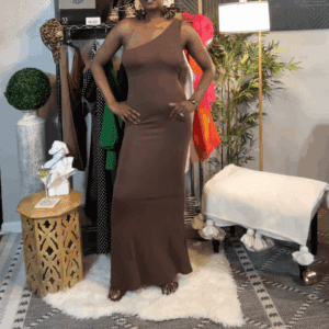A person stands indoors wearing the Long One Shoulder Back Out Dress in brown, surrounded by clothing, a plant, and decorative furniture.