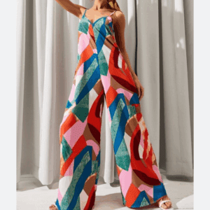 Indoors, a person wears the Spaghetti Strap Geometric Jumpsuit featuring wide legs and a bold red, blue, green, and pink geometric pattern. White curtains hang in the background.
