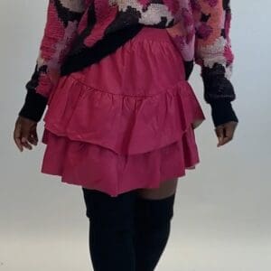A person wearing a Faux Leather Ruffle Skirt, a pink and black patterned sweater, and black knee-high boots stands against a plain background.