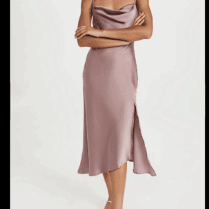 A woman wearing the Spaghetti Strap Dress—a sleeveless mauve satin midi with a cowl neckline and side slit—stands with her arms crossed, paired with strappy sandals.
